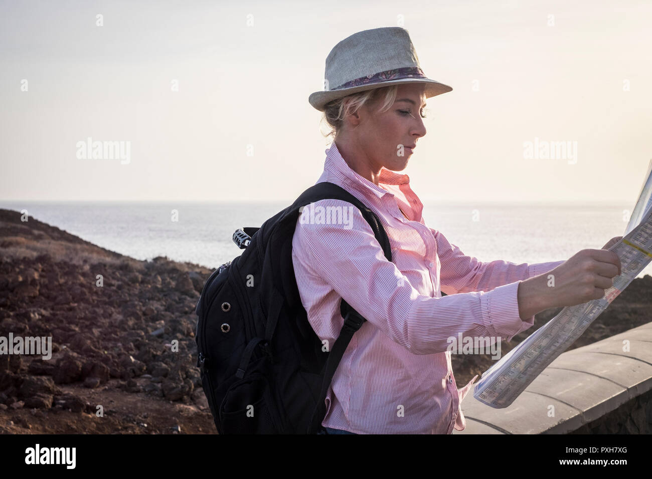 young blonde female traveler with black backpack read the map to choose the next destination. Coast and ocean in background. Outdoor travel concept Stock Photo