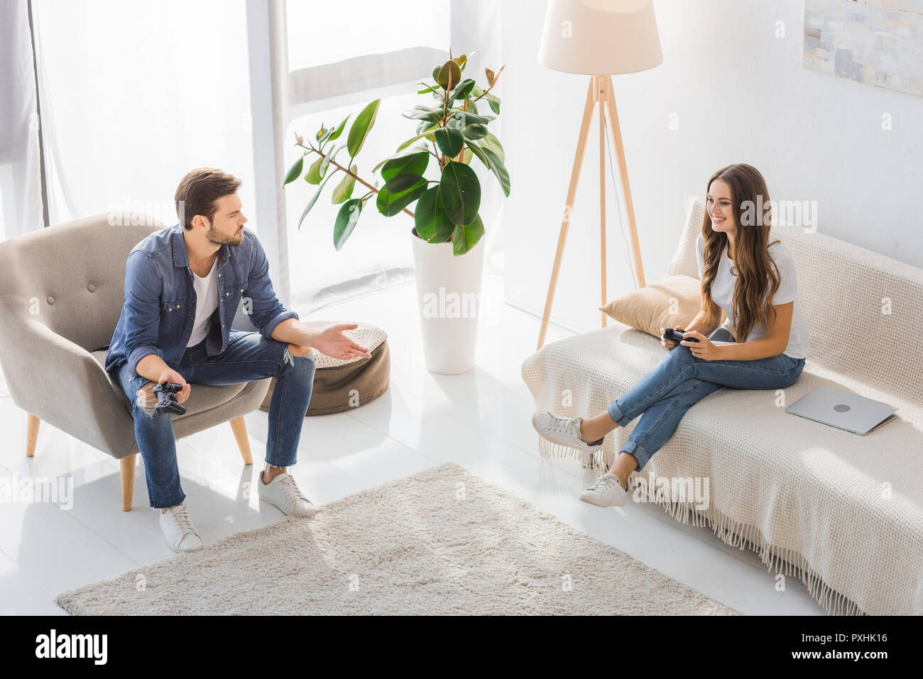 high angle view of young man on armchair doing dubium gesture while his smiling girlfriend sitting on sofa with joystick at home Stock Photo