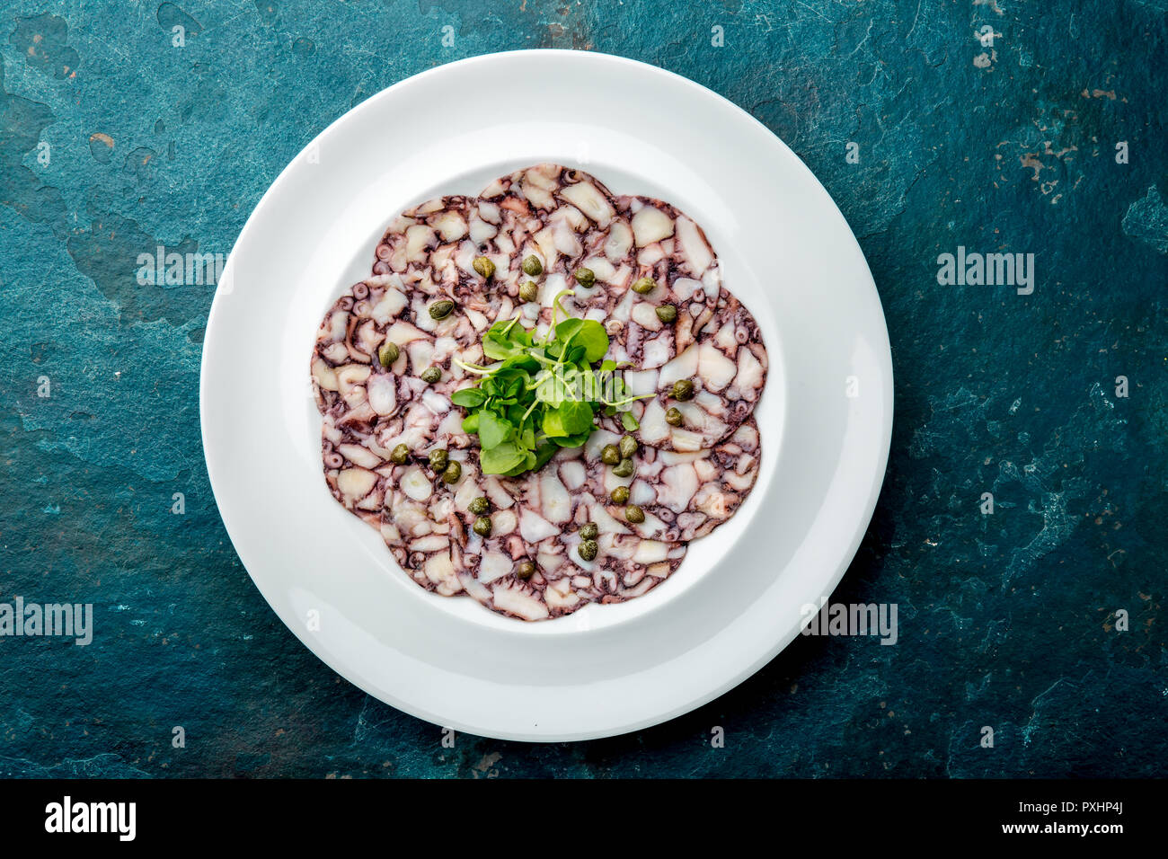OCTOPUS CARPACCIO. Seafood Raw octopus slices with olive oil, lemon and capers on white plate. Top view. blue background Stock Photo