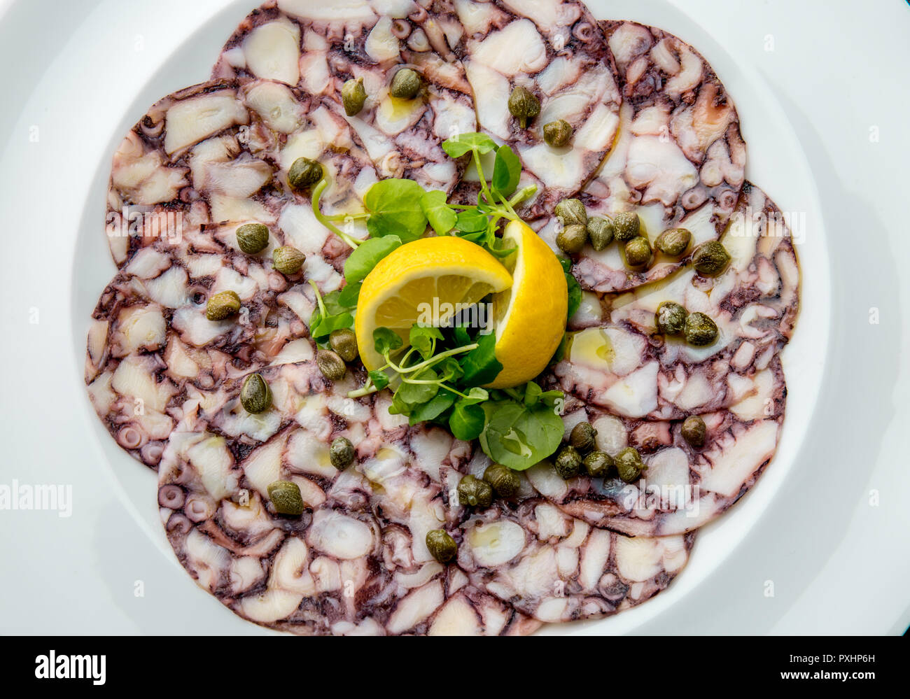 OCTOPUS CARPACCIO. Seafood Raw octopus slices with olive oil, lemon and capers on white plate. Top view. Gray stone background Stock Photo