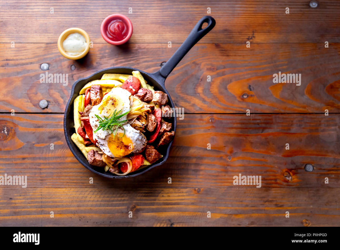 CHILEAN FOOD. CHORRILLANA - french fries topped with beef sliced, tipical sausages chorrisos, fried onion and eggs served in cast iron pan with sauces Stock Photo
