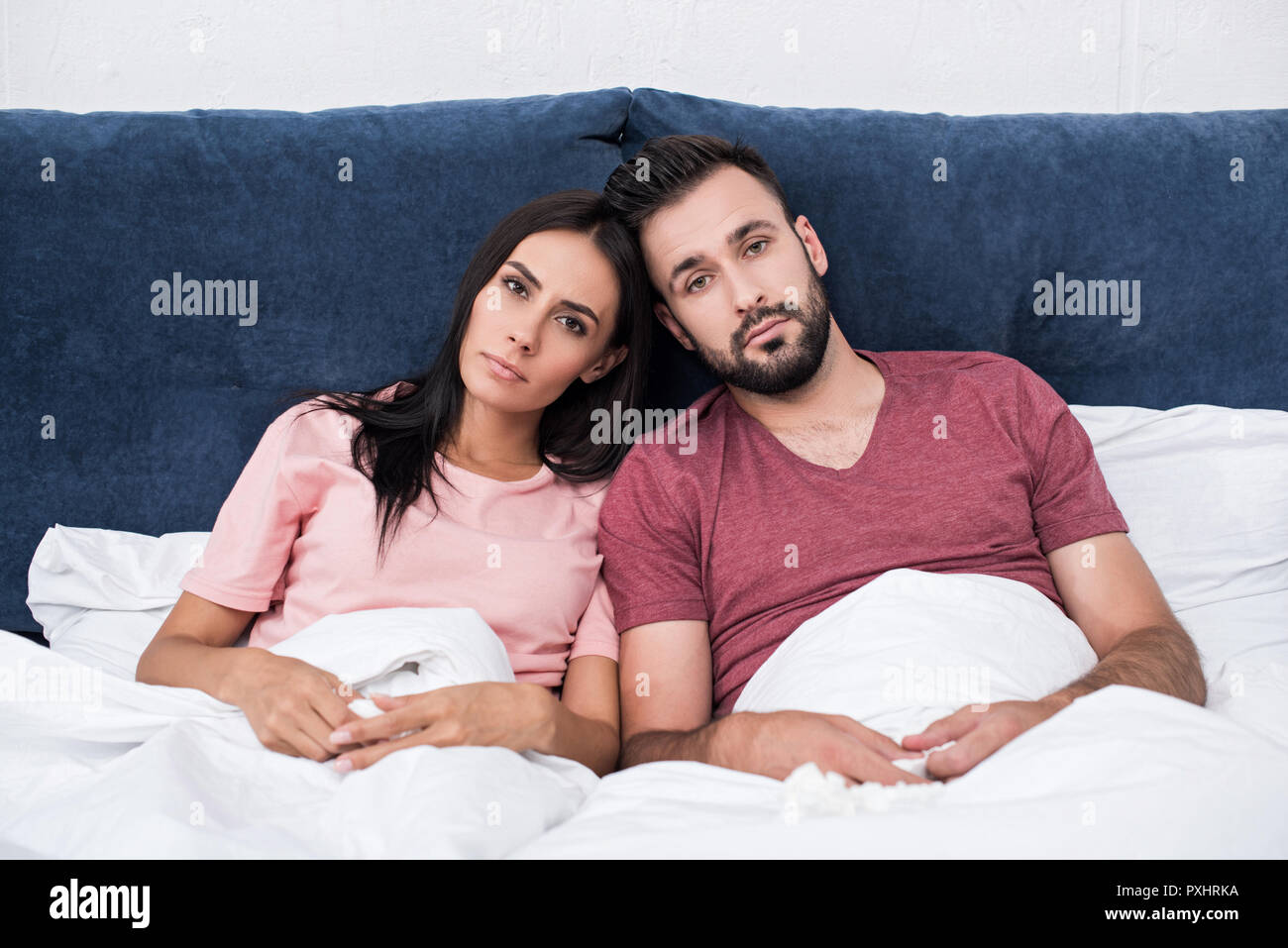unemotional young couple leaning at each other while sitting in bed and looking at camera Stock Photo