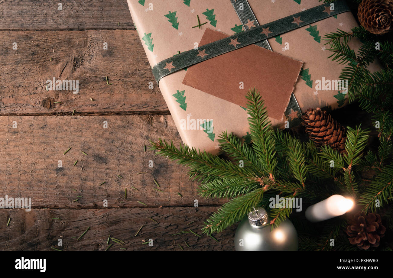 Green fir twigs, pine cones, and Christmas balls, near a gift wrapped in classic brown paper with trees, and a lit candle, on a vintage wooden table. Stock Photo