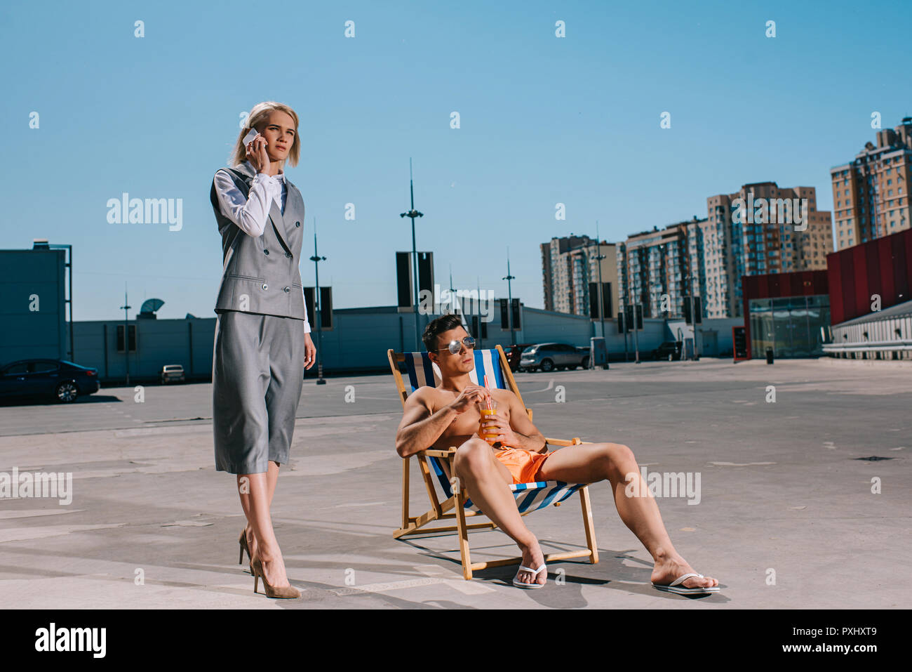 attractive young man relaxing on sun lounger while his lady boss in suit next to him and talking by phone on parking Stock Photo