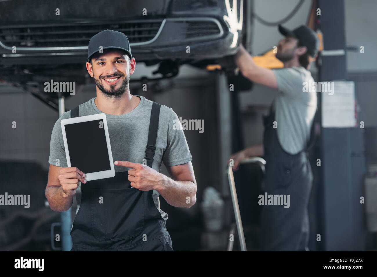 smiling mechanic showing digital tablet with blank screen, while colleague working in workshop behind Stock Photo