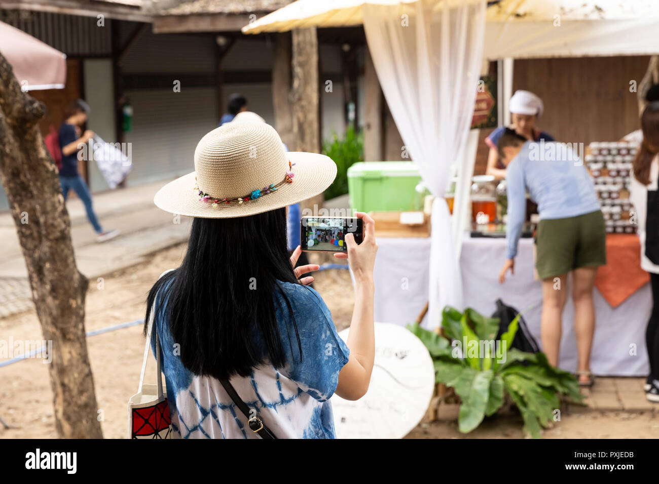 Woman in hat pictured from back holding smartphone taking photo at JingJai Farmer's Market, Chiang Mai, Thailand Stock Photo