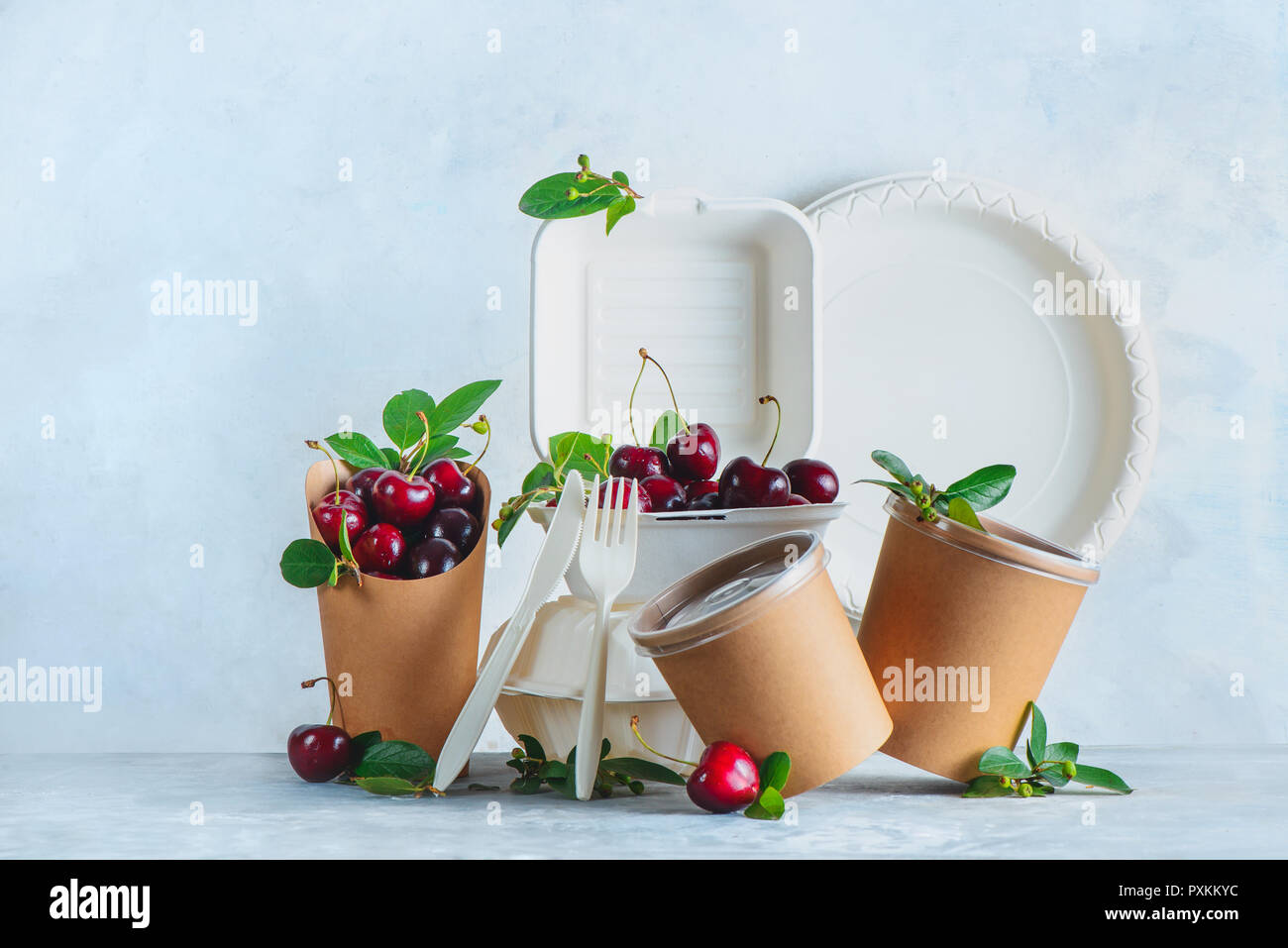 Catering disposables, cups, plates and containers with cherries. Eco-friendly food packaging on a neutral gray background with copy space. Preserving  Stock Photo