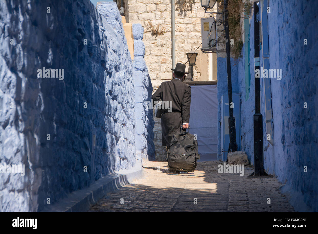 Ultraorthodox jew walking through the blue painted streets of Safed (Tsfat), Israel, with a wheeled suitcase. Stock Photo