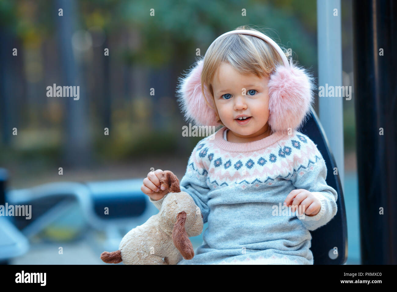 Portrait of sweet blond little baby girl in beautiful warm dress and winter faux fur ear muffs at playground Stock Photo
