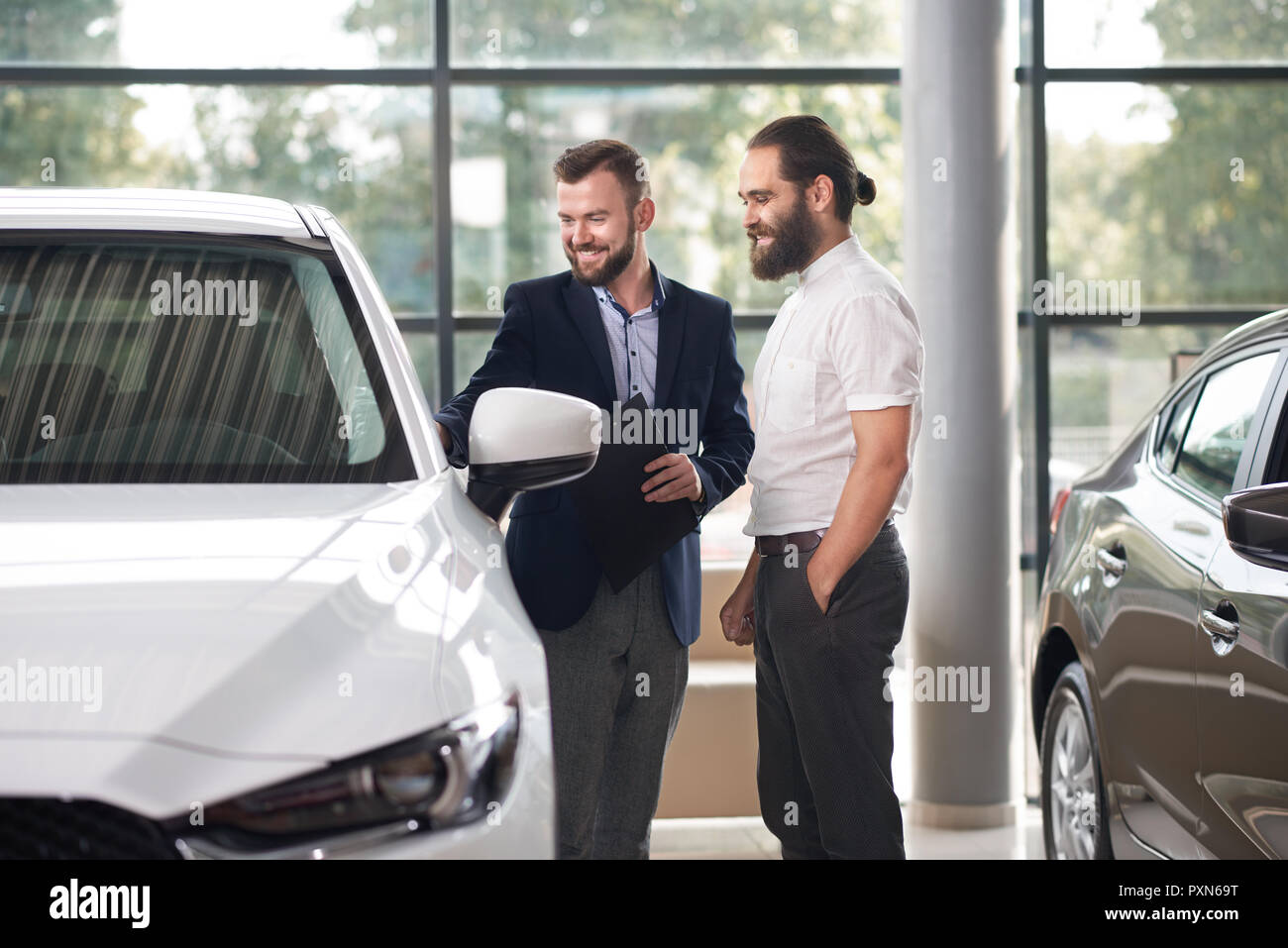 Manager holding folder and showing to customer of car center white automobile. Men standing near auto, looking at it and smiling. Car dealer wearing in dark blue jacket, buyer in white shirt. Stock Photo