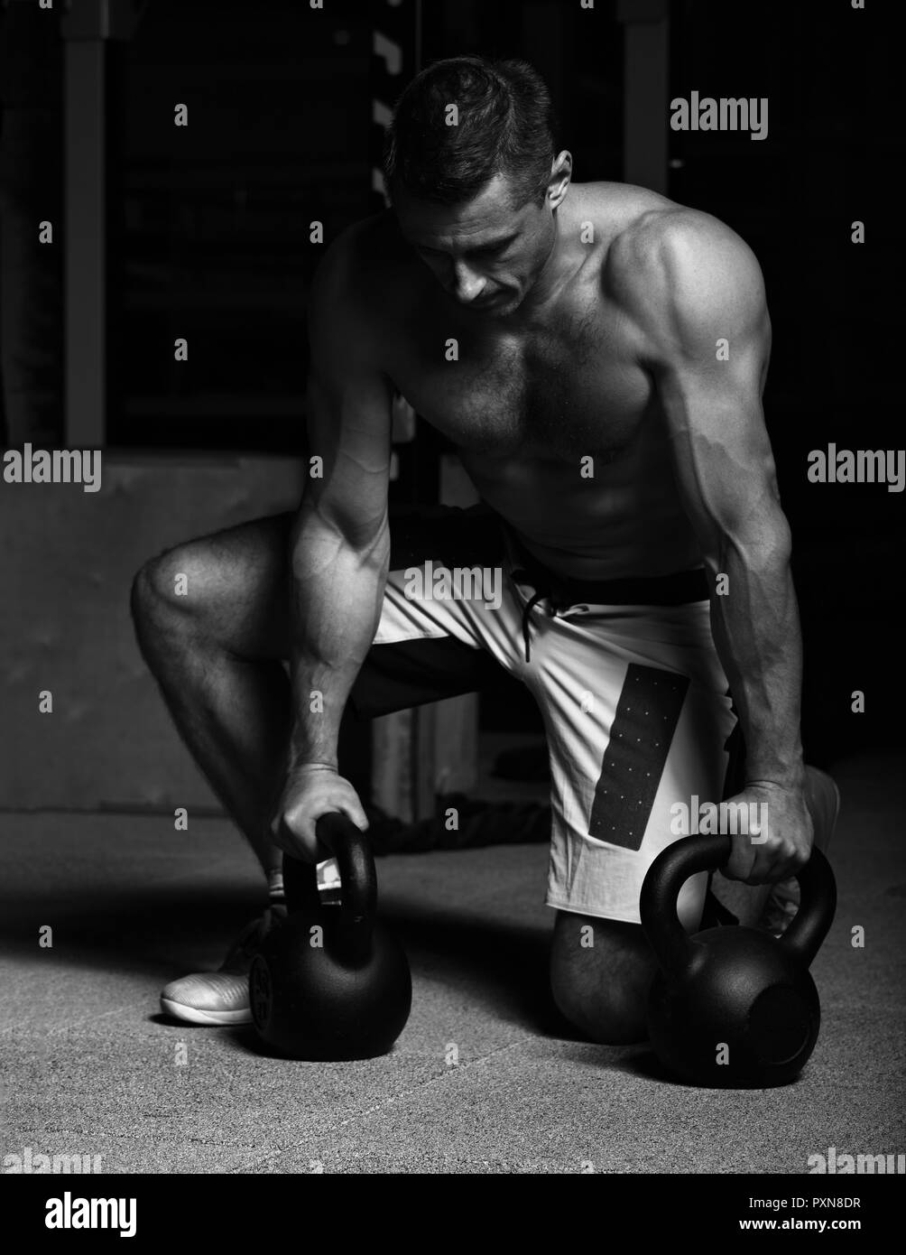 Handsome serious strong brutal man sitting and holding kettlebells on dark sport club background. Training time. Contrast portrait. Black and white Stock Photo