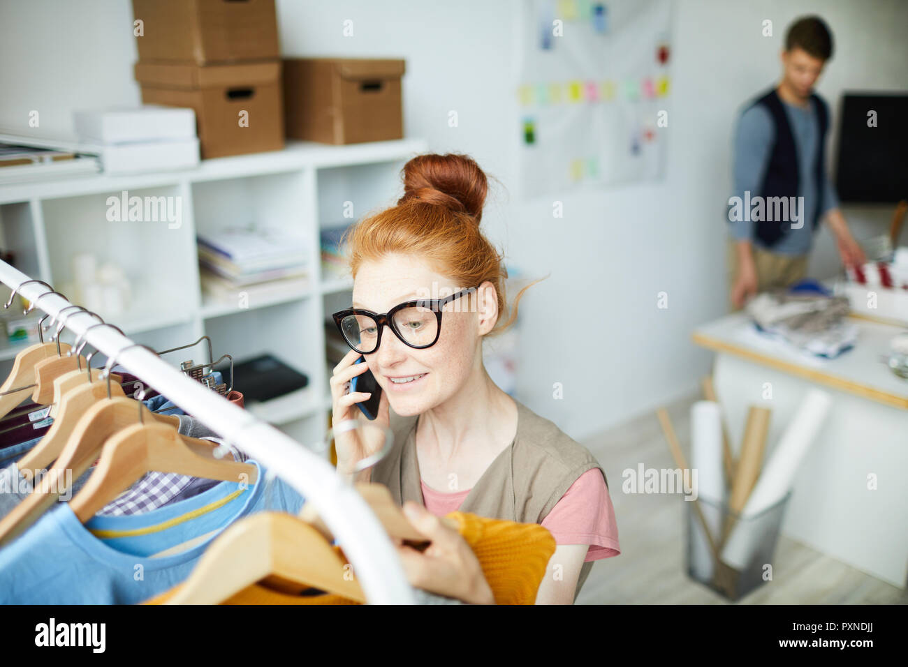 Young female designer in eyeglasses looking at clothes on rack and consulting on the phone Stock Photo