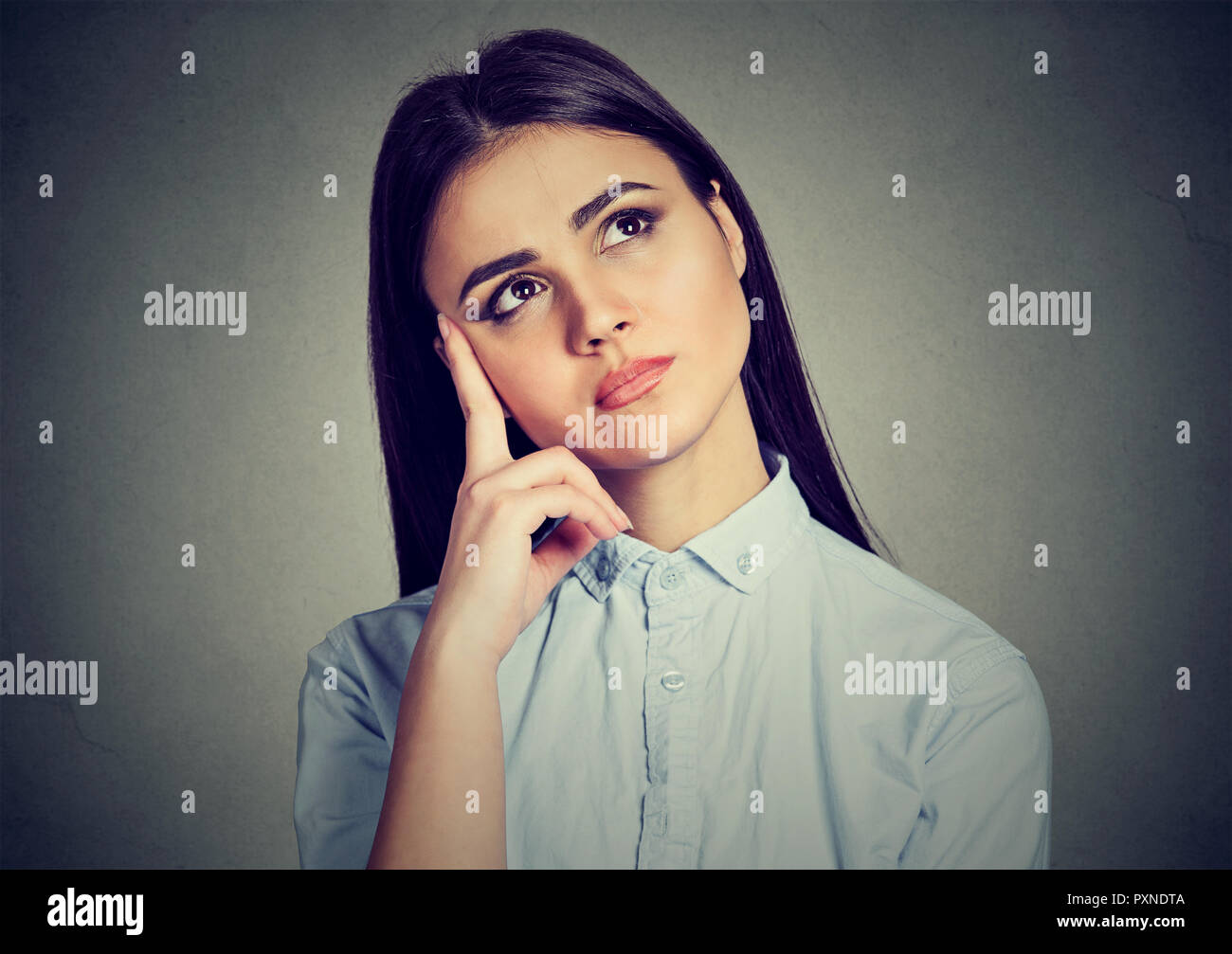 Beautiful young woman in blue shirt touching face in contemplation looking away and up thinking Stock Photo