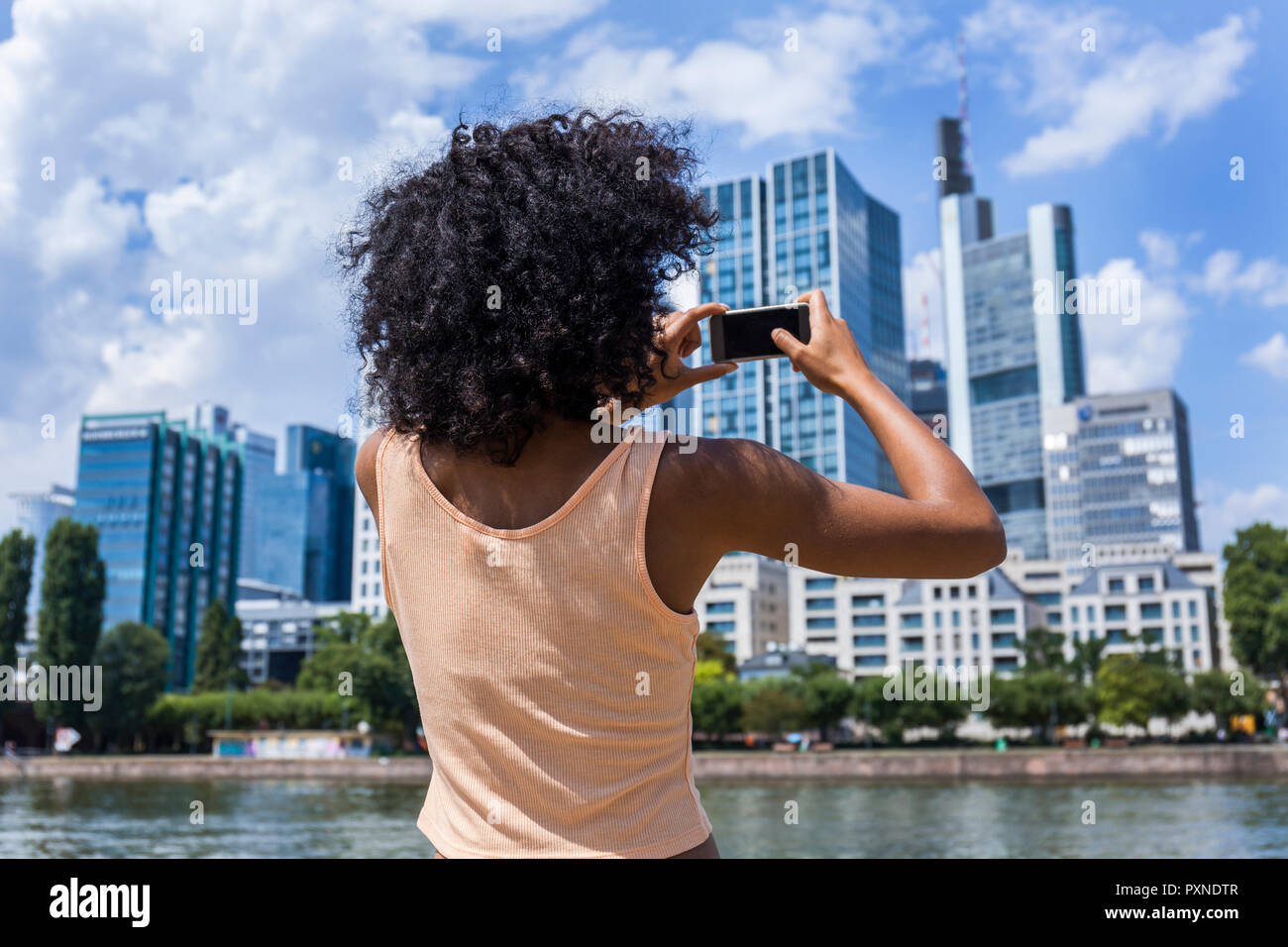 Germany, Frankfurt, back view of young woman with curly hair taking photo with smartphone Stock Photo