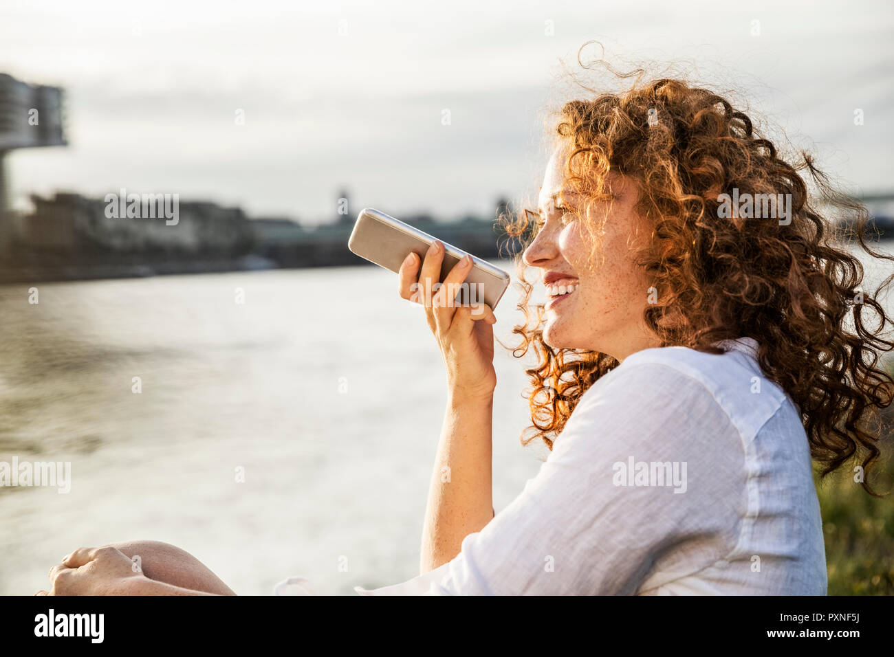 Germany, Cologne, portrait of laughing young woman on the phone sitting at riverside in the evening Stock Photo