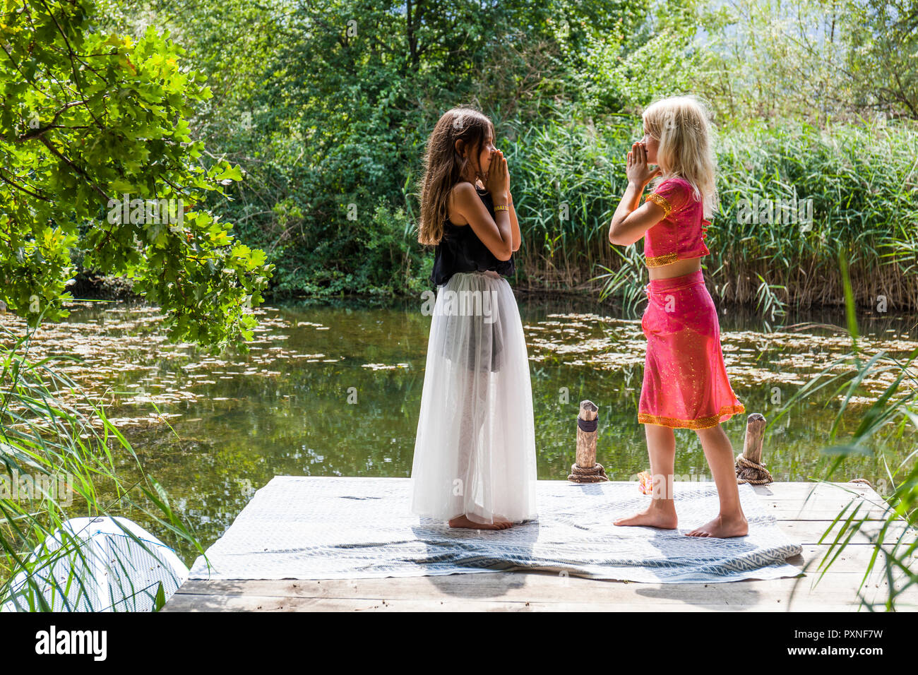 Two girls standing face to face on jetty at a pond in fancy dresses Stock Photo