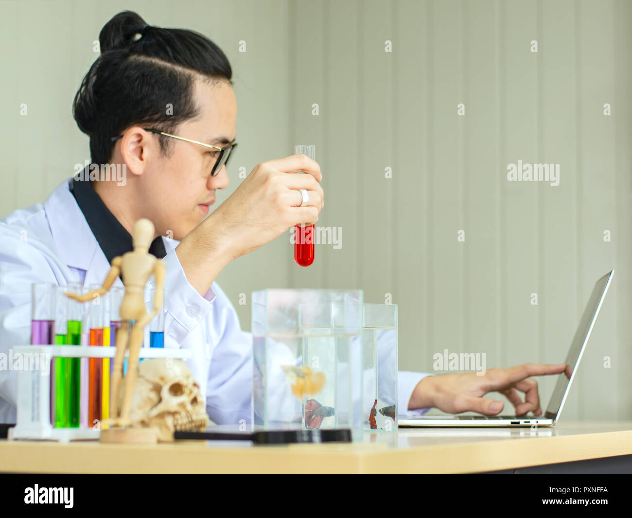 Asian scientist wearing eyeglassess holding tube contain red liquid witht Siam fighting fish in tank while recording testing result in notebook Stock Photo