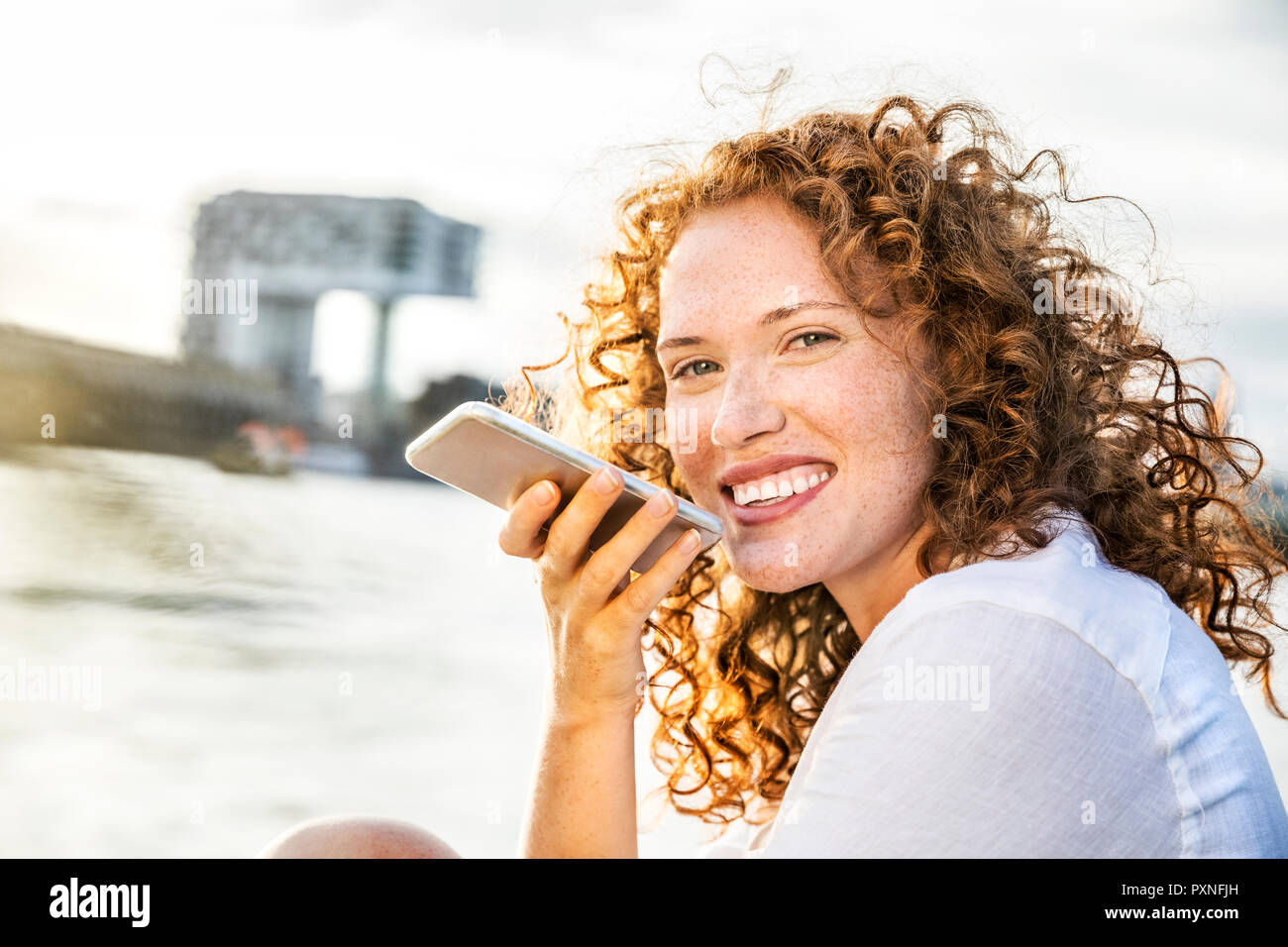 Germany, Cologne, portrait of happy  young woman on the phone sitting at riverside in the evening Stock Photo
