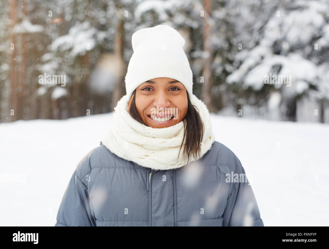 Cheerful excited young mixed race girl in white hat and scarf standing under falling snow and looking at camera in winter forest Stock Photo