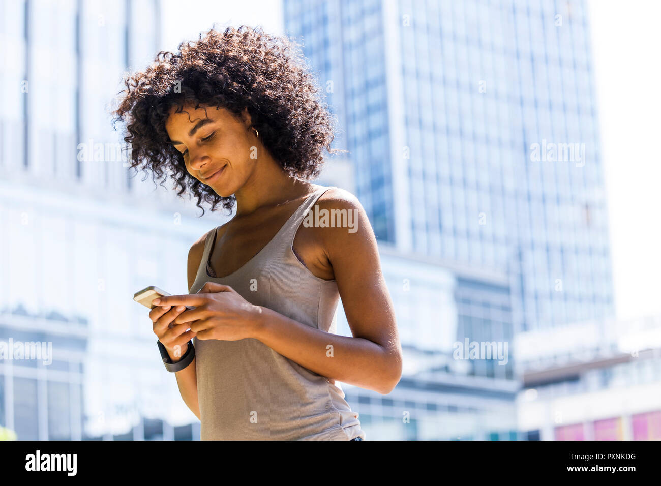 Germany, Frankfurt, smiling young woman with curly hair looking at cell phone in front of skyscrapers Stock Photo