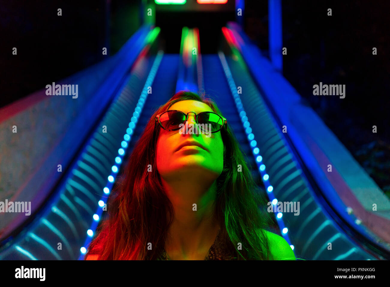 Portrait of illuminated young woman wearing sunglasses in front of blue lighted escalator Stock Photo