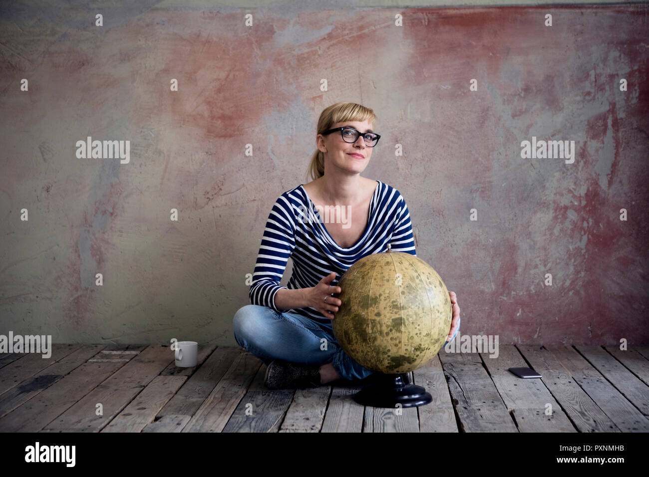 Smiling woman sitting on wooden floor in an unrenovated room with an old globe Stock Photo