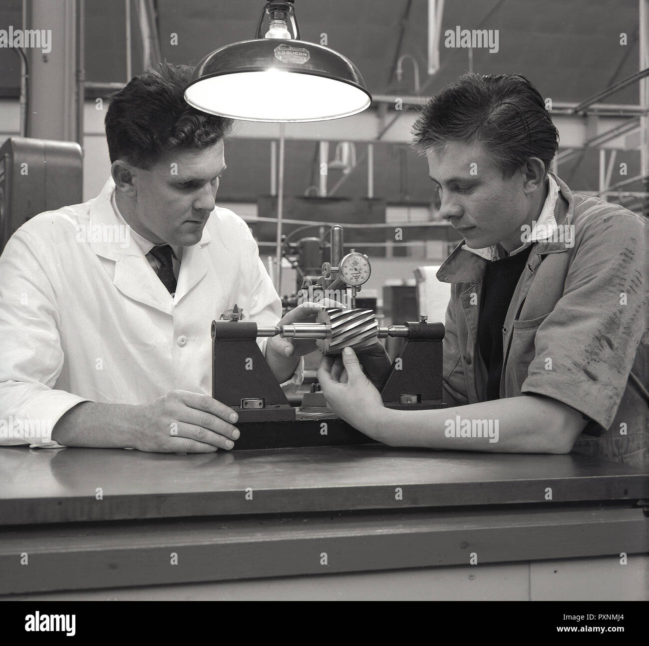 1962, historial, inside a facory, a young male aviation engineering apprentice sits with his white-coated male engineer teacher or mentor, England, UK. Stock Photo