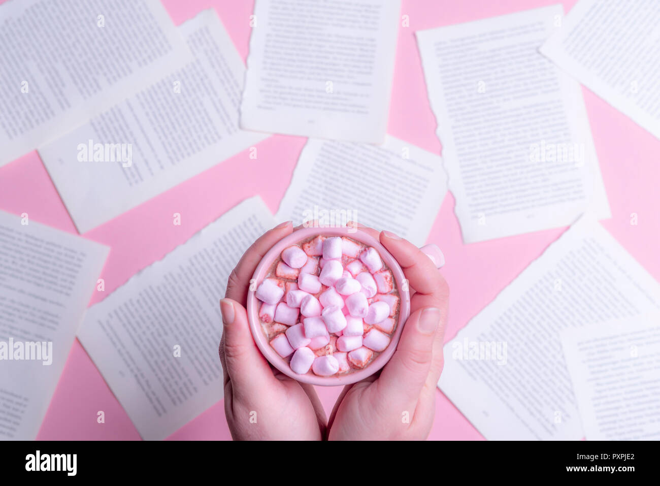 Woman holding, with both hands, a pink cup of hot chocolate with pink mini marshmallows, above a table full of book pages. Stock Photo