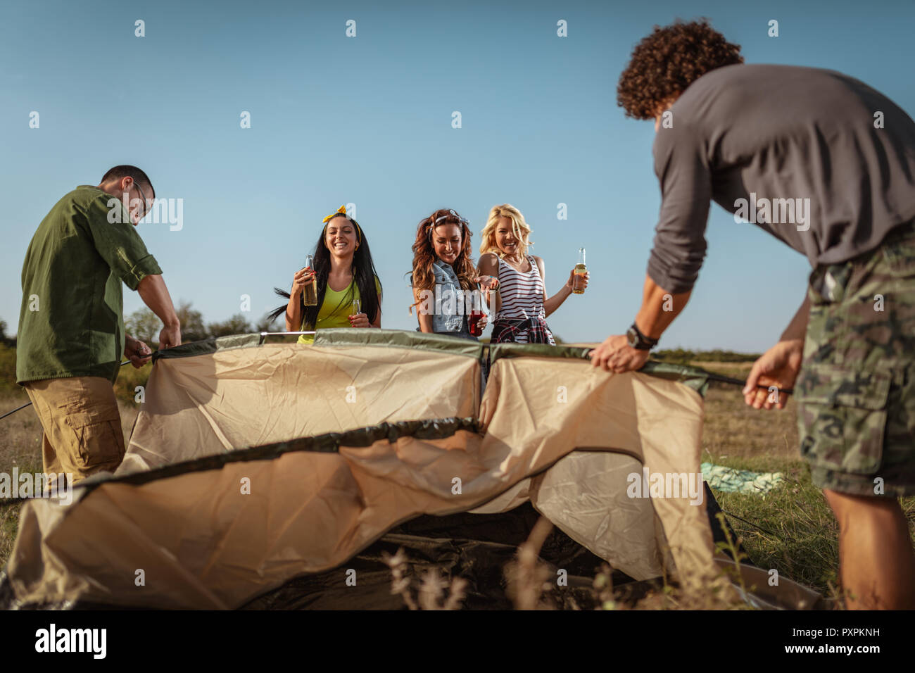 The young happy friends are preparing for camping. They're installing a tent on a suitable place in a meadow and their girlfriends are offering the be Stock Photo