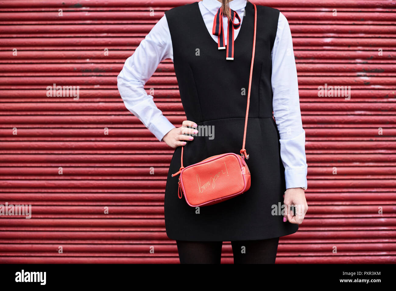 Fashionable woman with red handbag wearing black dress standing in front of red roller shutter Stock Photo