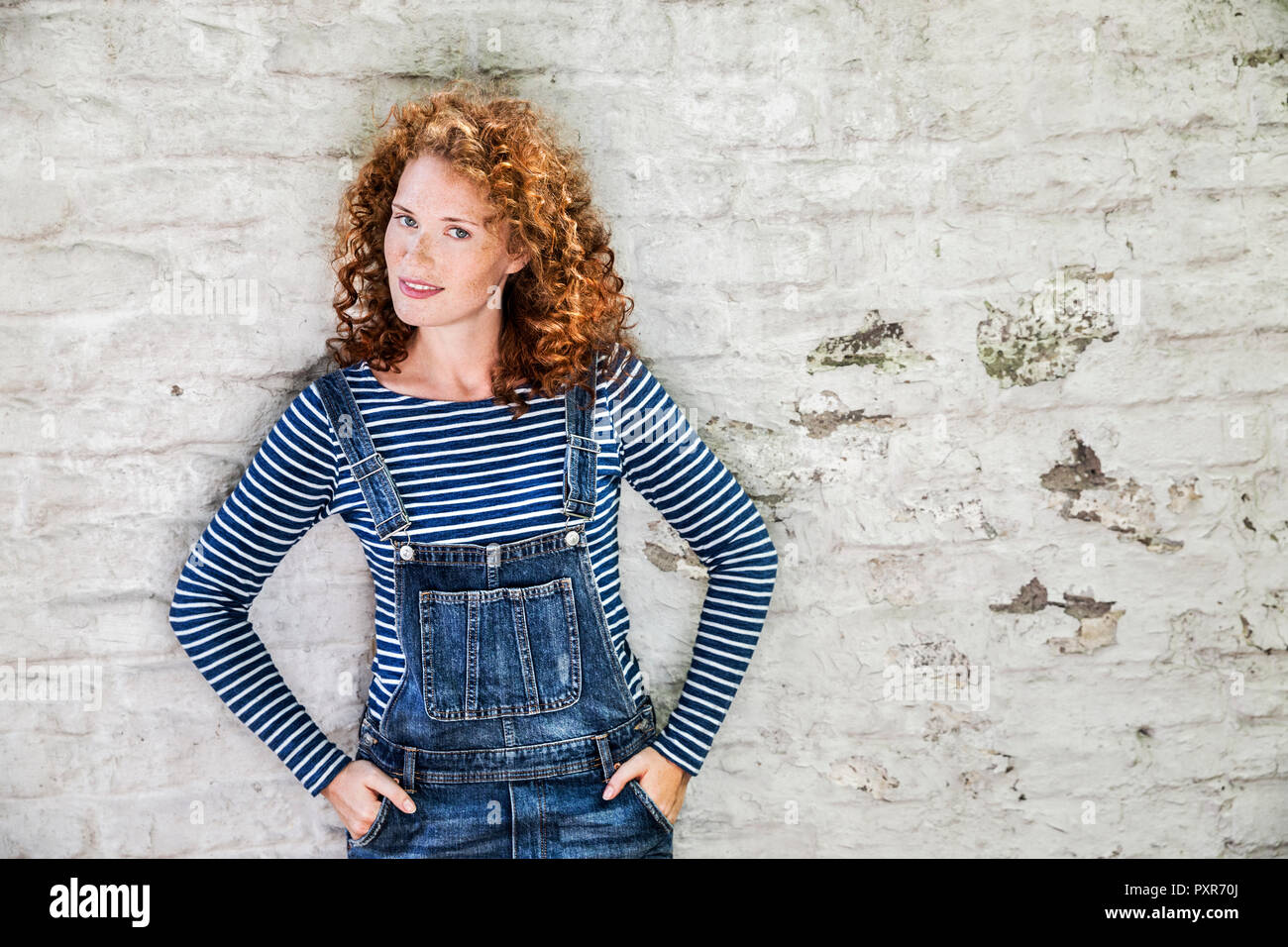 Portrait of smiling young woman wearing denim dungarees leaning against white brick wall Stock Photo