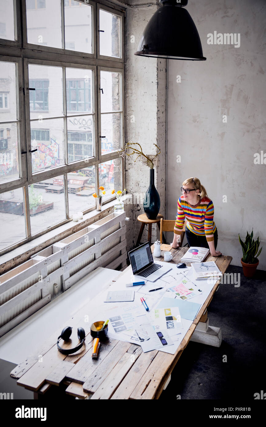 Woman standing at desk in a loft looking through window Stock Photo