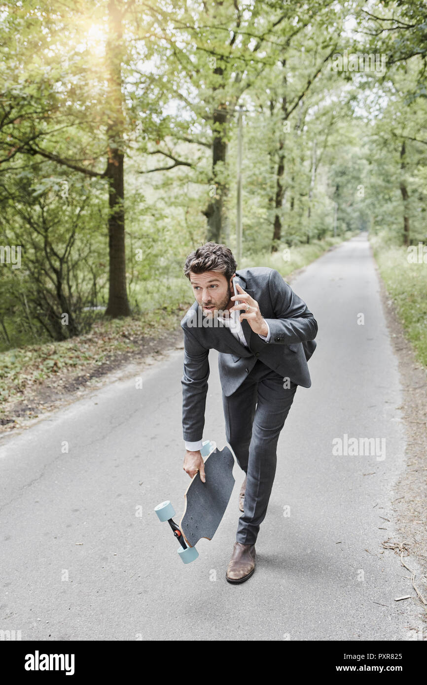Businessman walking with skateboard and smartphone on rural road Stock Photo