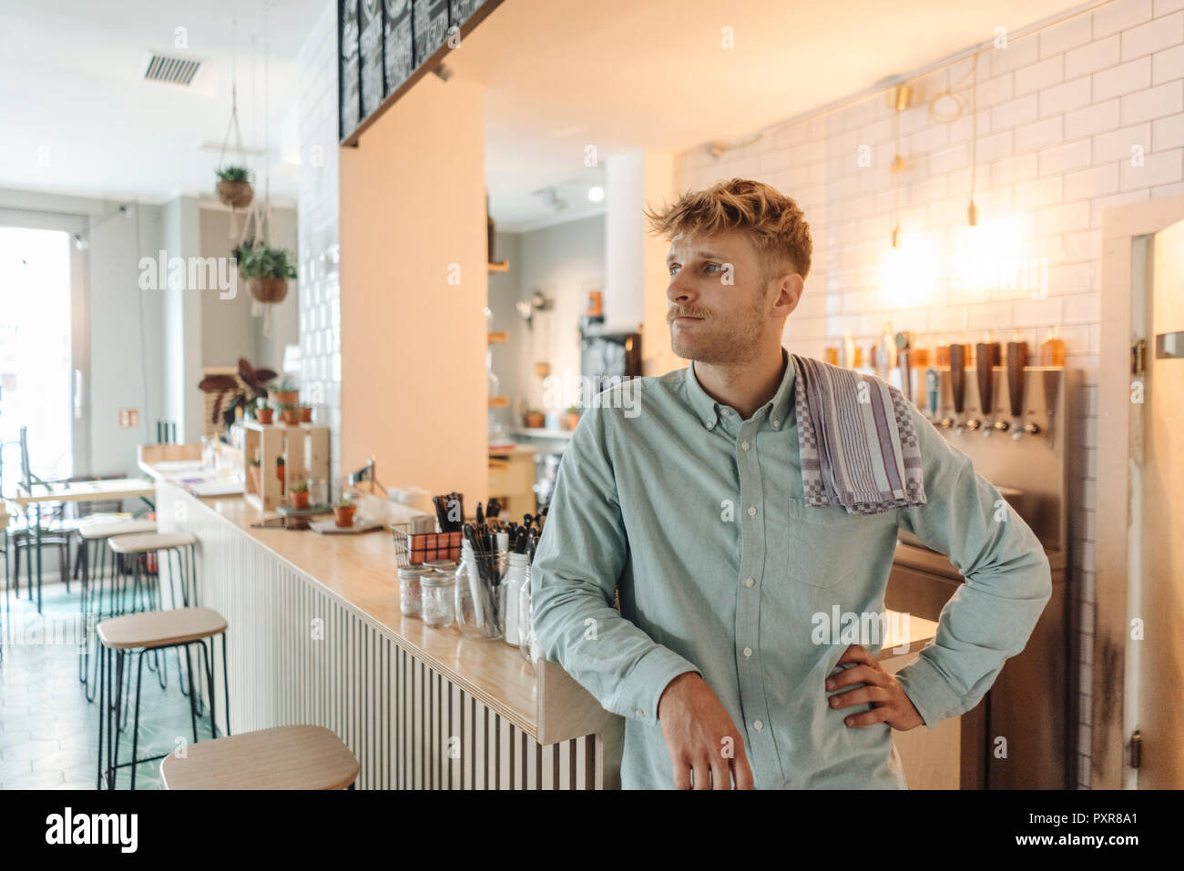 Young man working in his start-up cafe, standing at bar, daydreaming Stock Photo