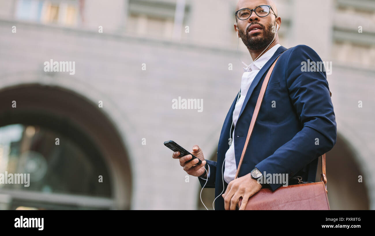 Low angle view of young african businessman with bag walking outdoor on street listening to music from mobile phone. Handsome businessman using smartp Stock Photo