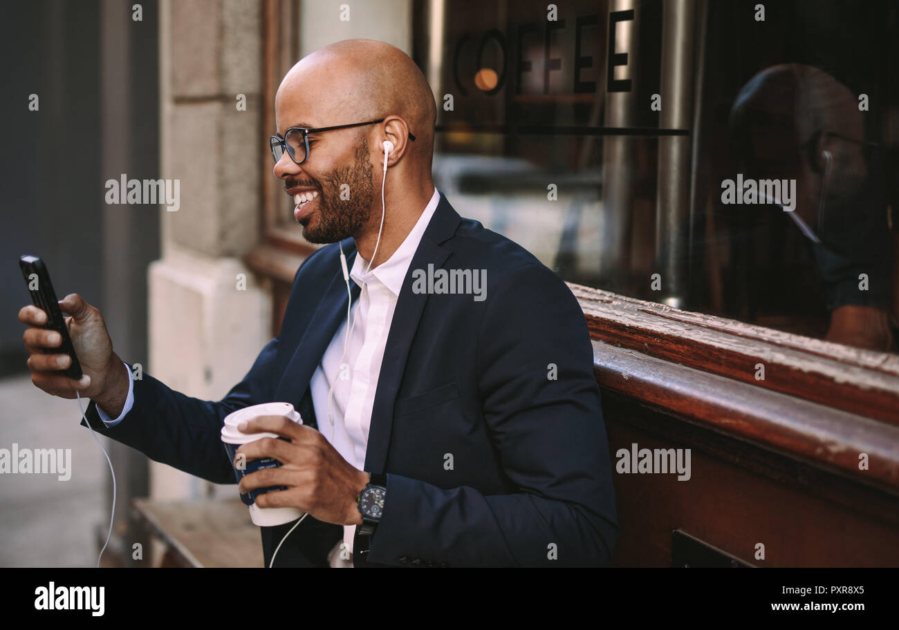 Happy young african businessman commuting with a smartphone. African businessman with earphones making a video call sitting outdoors. Stock Photo
