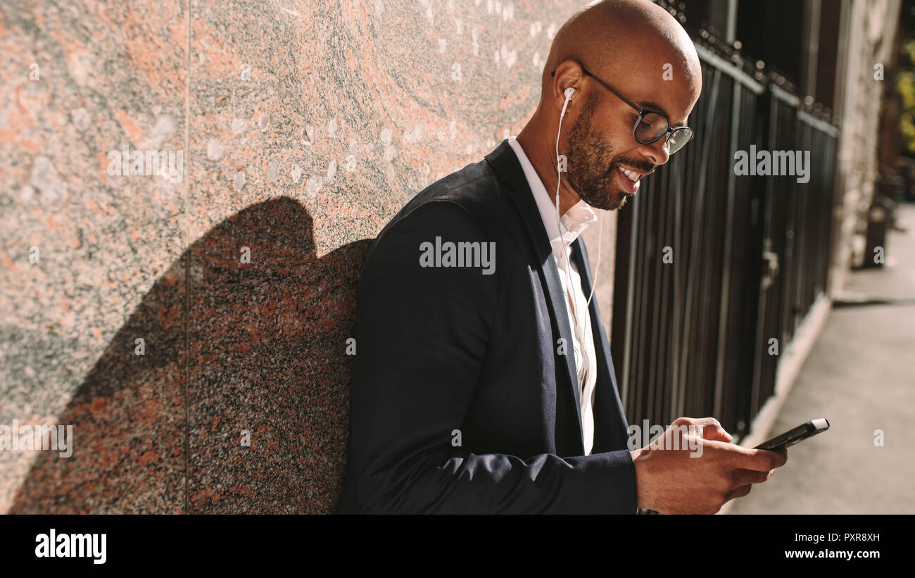Smiling young businessman leaning on a wall while standing outdoors using mobile phone. Man in suit wearing earphones looking at his smart phone. Stock Photo
