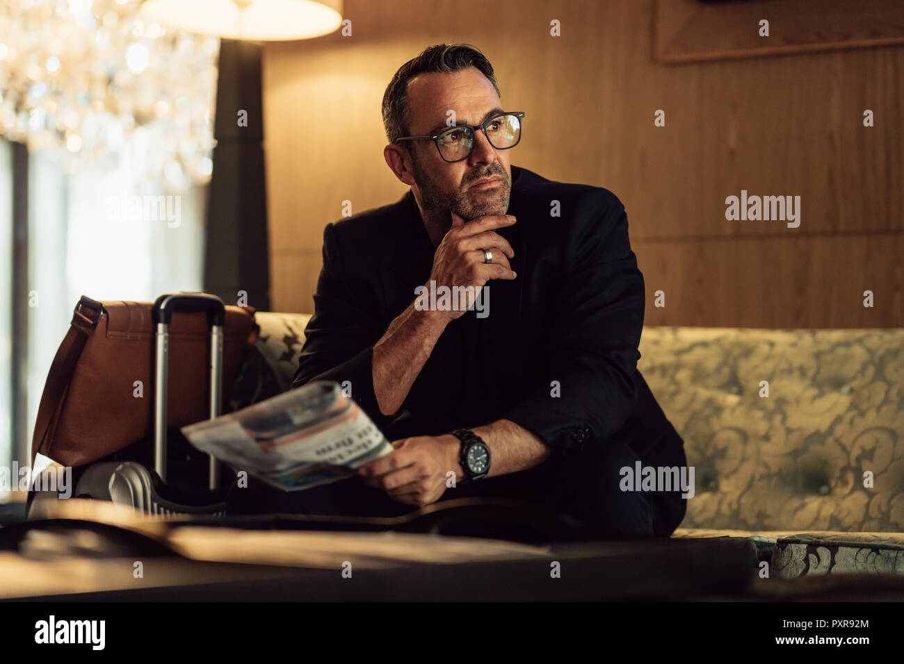 Portrait of thoughtful mature businessman sitting in hotel lobby with his luggage and looking away. Mature CEO waiting in hotel lobby holding newspape Stock Photo