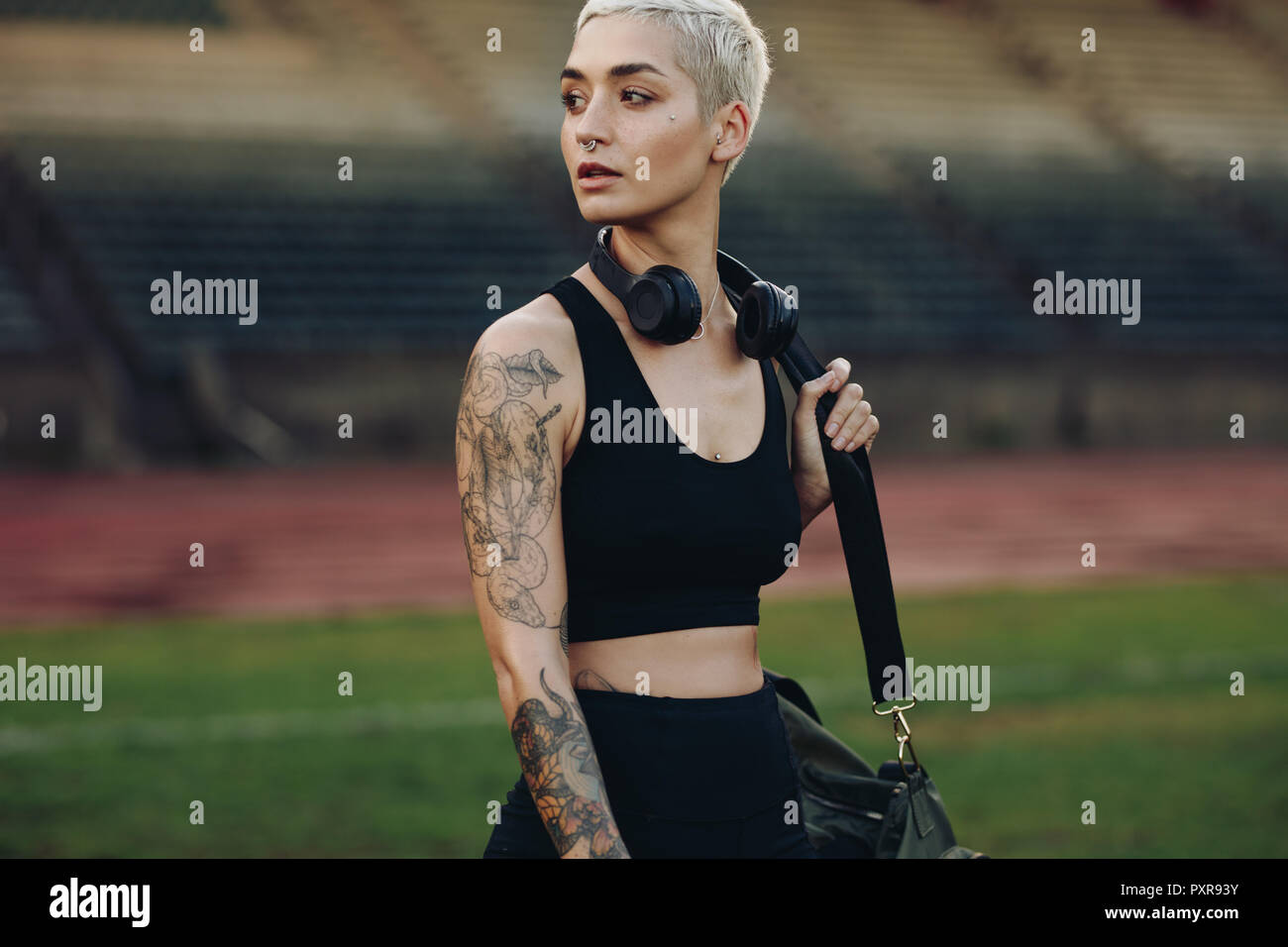 Female athlete walking in a track and field stadium carrying her gym bag. Woman in fitness wear with a wireless headphone on neck looking away. Stock Photo