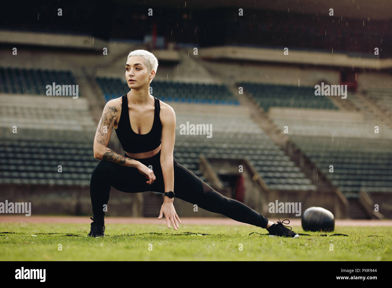 Woman athlete doing stretching exercises in a stadium with a medicine ball by her side. Female athlete doing stretching workout in a track and field s Stock Photo