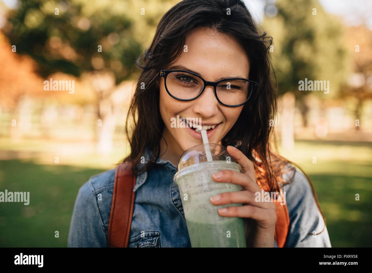 Cheerful woman at park drinking juice and winking. Woman in eyeglasses drinking smoothie and winking an eye. Stock Photo