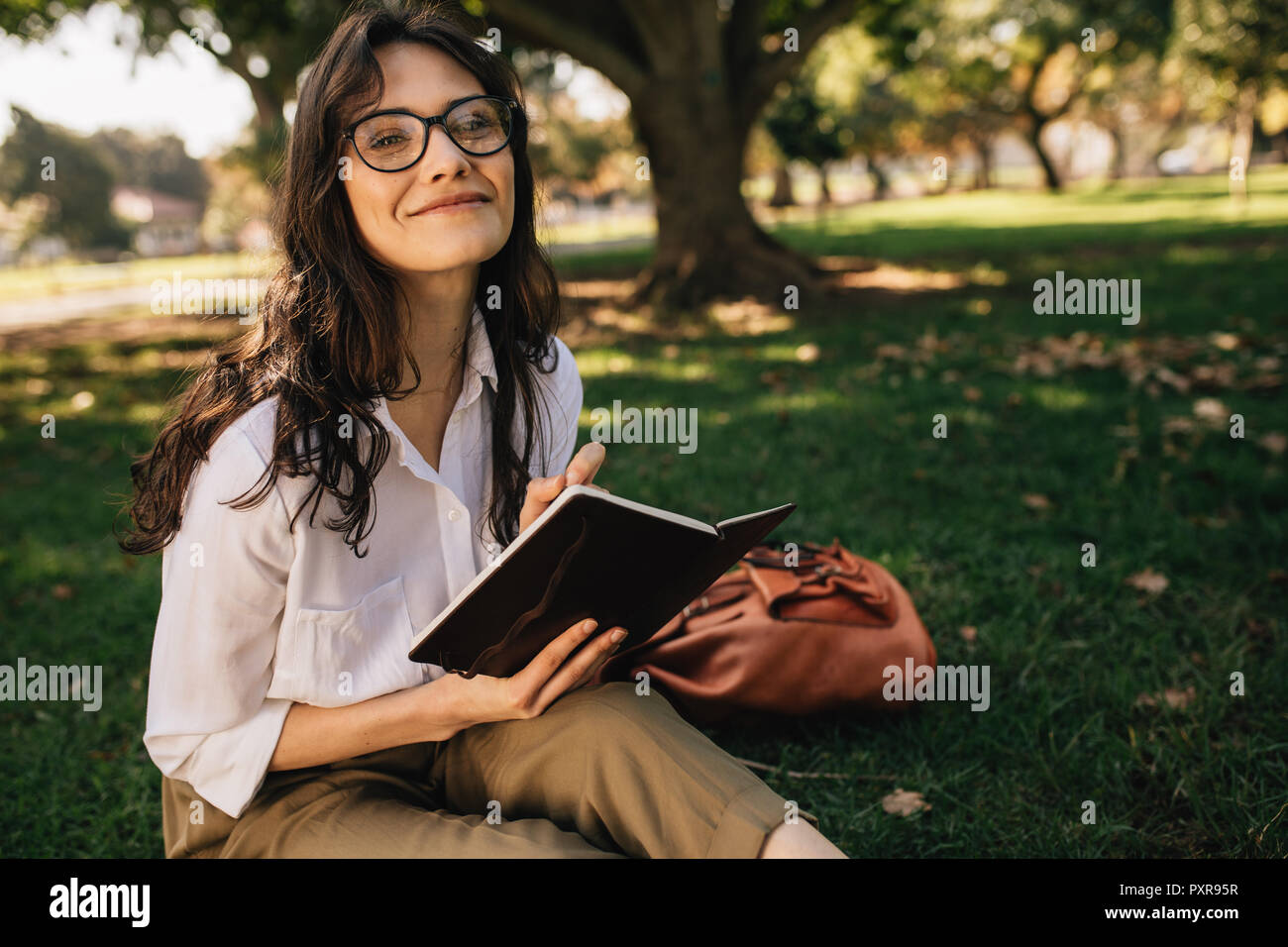 Pretty woman writing in book at park. Female wearing eyeglasses sitting at park with a book. Stock Photo