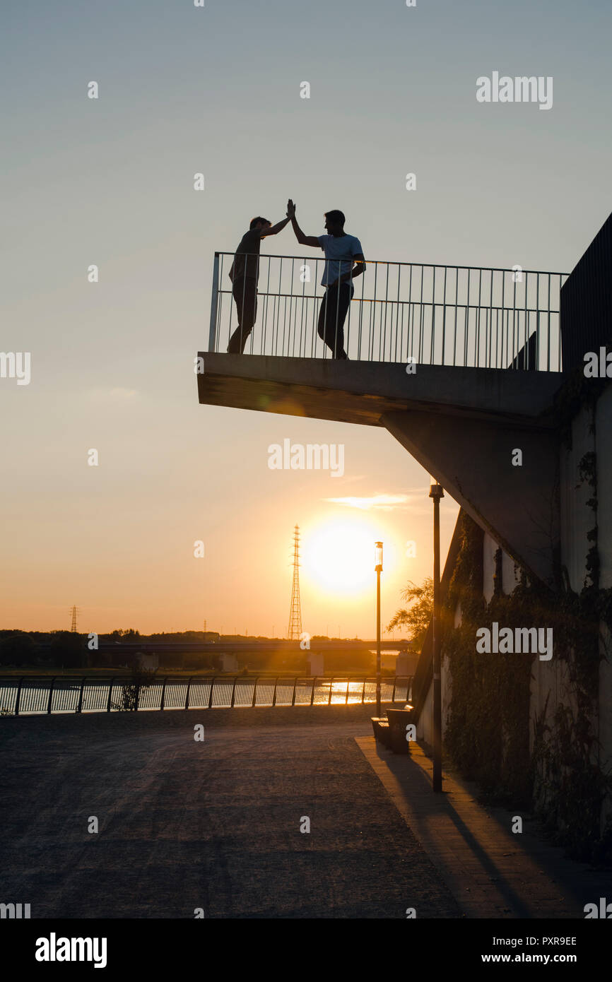 Two friends high-fiving at sunset, standing on observation platform Stock Photo