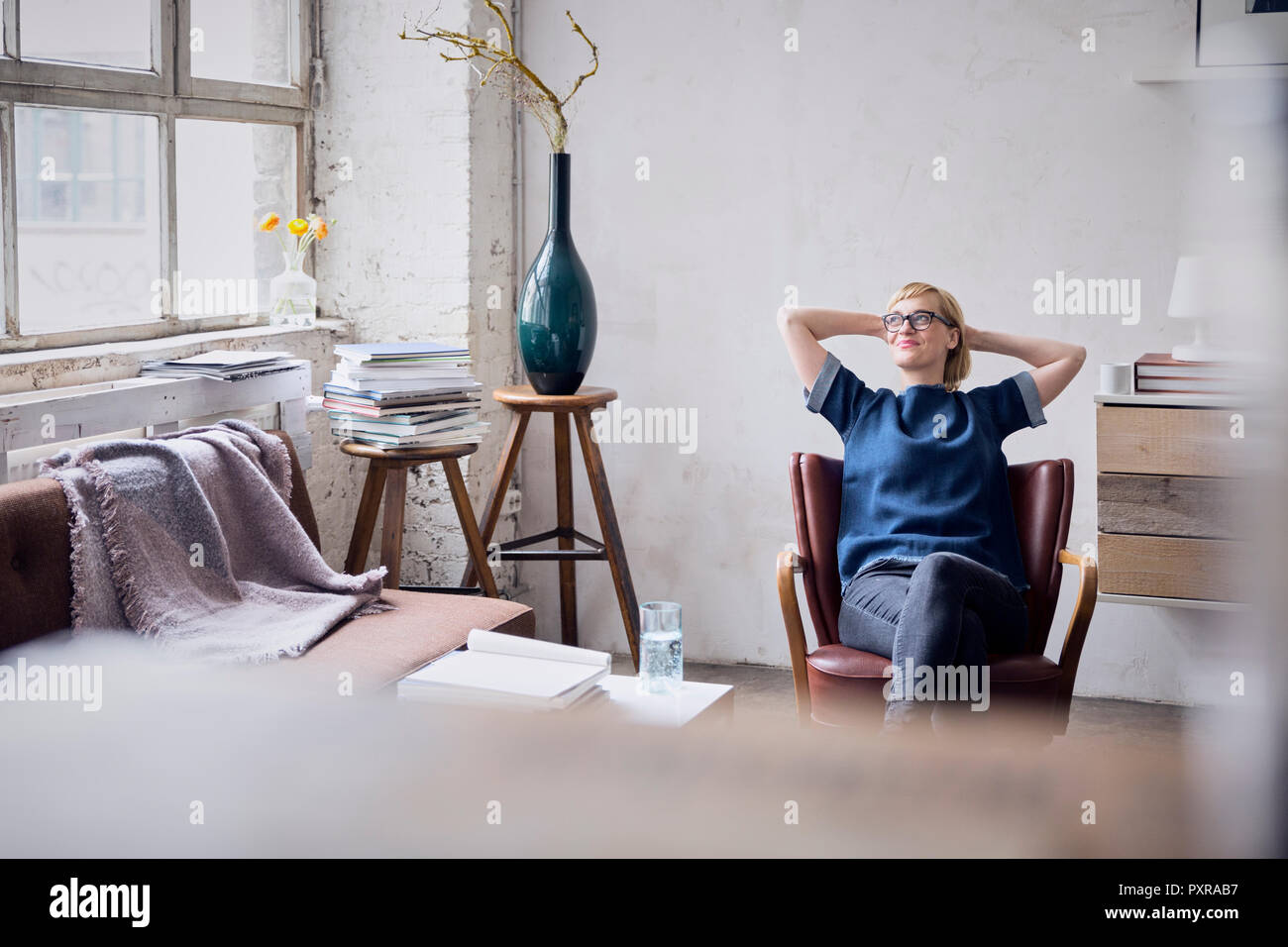 Smiling woman sitting on arm chair in loft looking through window Stock Photo
