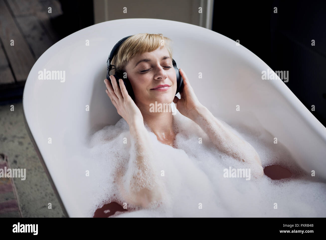 Blond woman taking bubble bath in a loft listenung music with headphones Stock Photo
