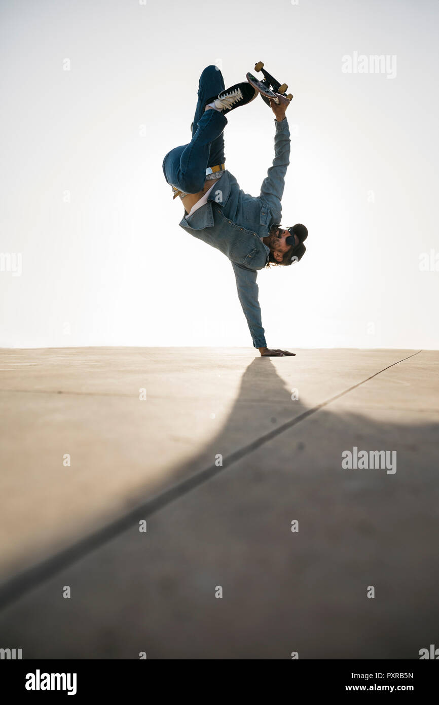 Stylish man in denim outfit showing trick with skate in handstand Stock Photo