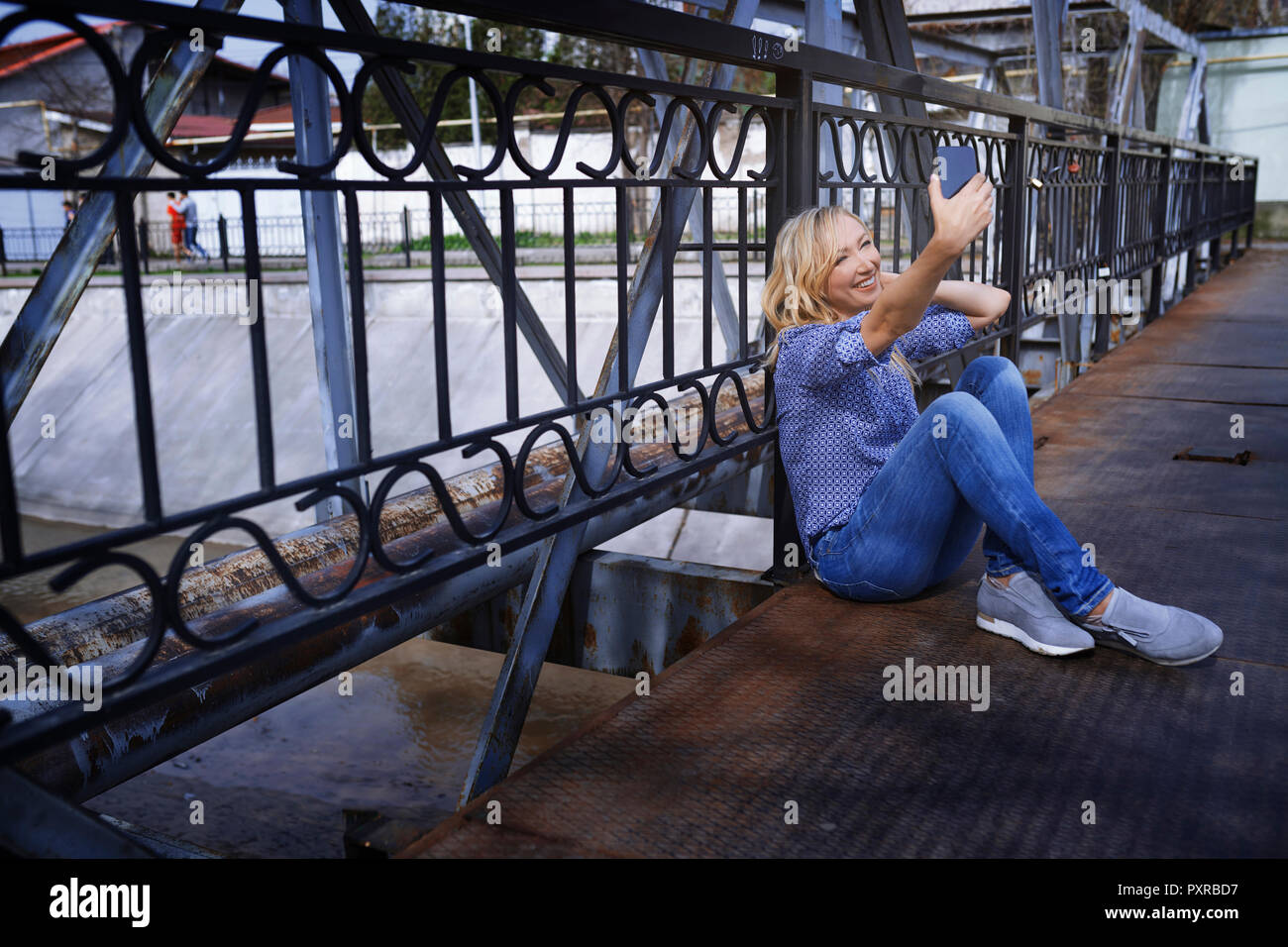 Blond woman sitting on bridge, taking selfie Stock Photo