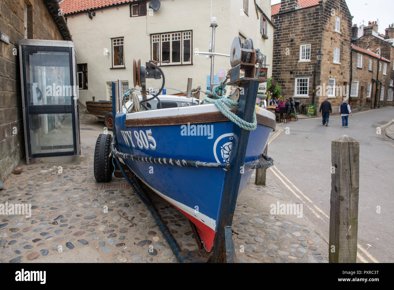 Tied down to a metal trailer, a small blue boat small rests on shore in Robin Hood's Bay, Yorkshire, UK Stock Photo