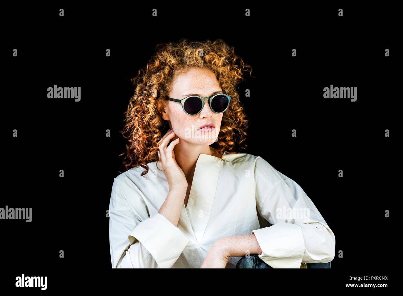 Portrait of stylish young woman with curly red hair wearing sunglasses in front of black background Stock Photo
