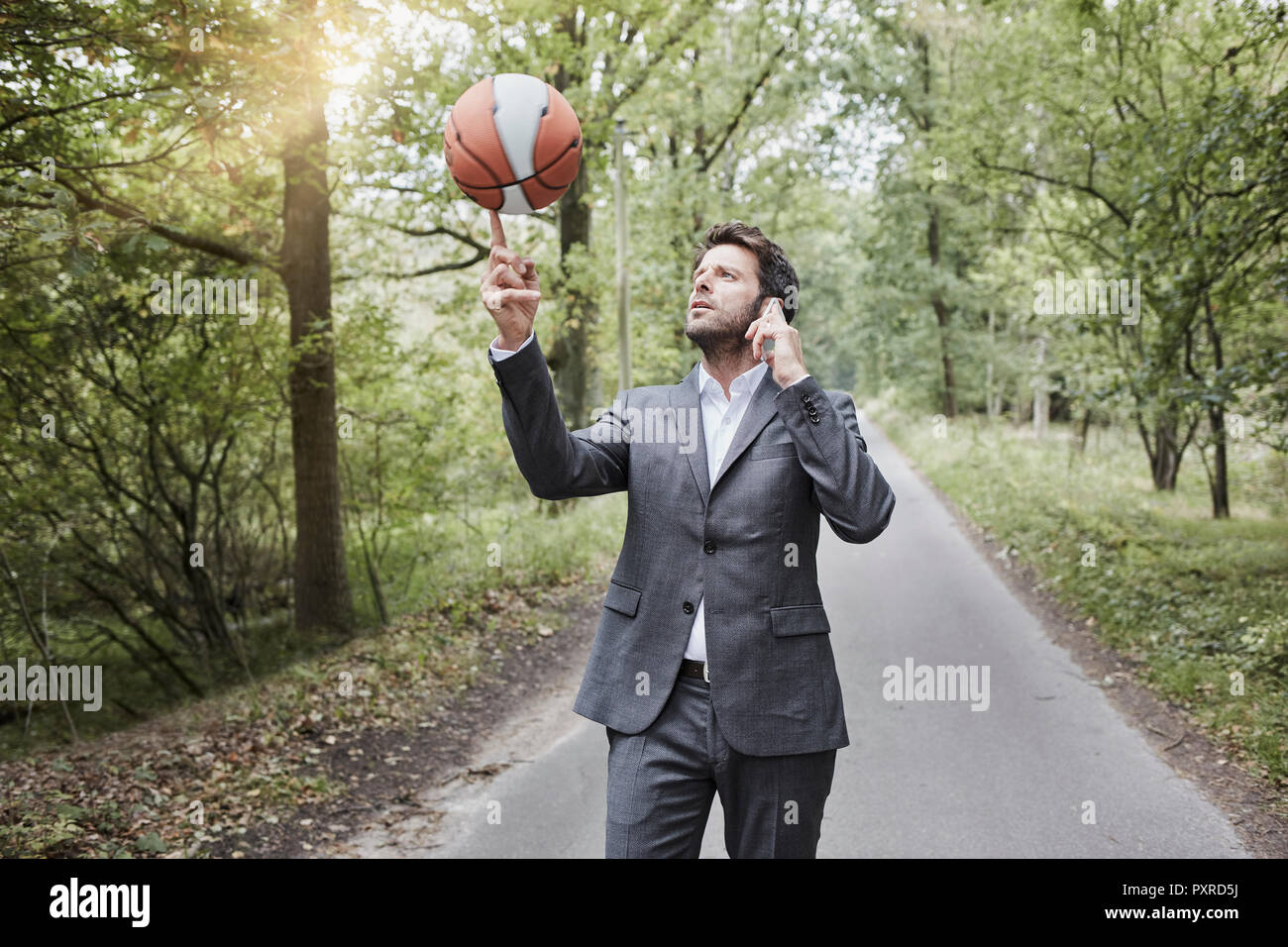 Businessman balancing basketball and talking on smartphone on rural road Stock Photo